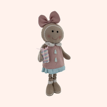 A plush gingerbread doll wearing a pink dress with a blue skirt, a pink bow on its head, and holding a small wrapped gift.