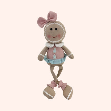 A gingerbread-themed fabric doll with a round tan head, pink bow, pink dress, light blue skirt, and pink booties, shown on a plain background.