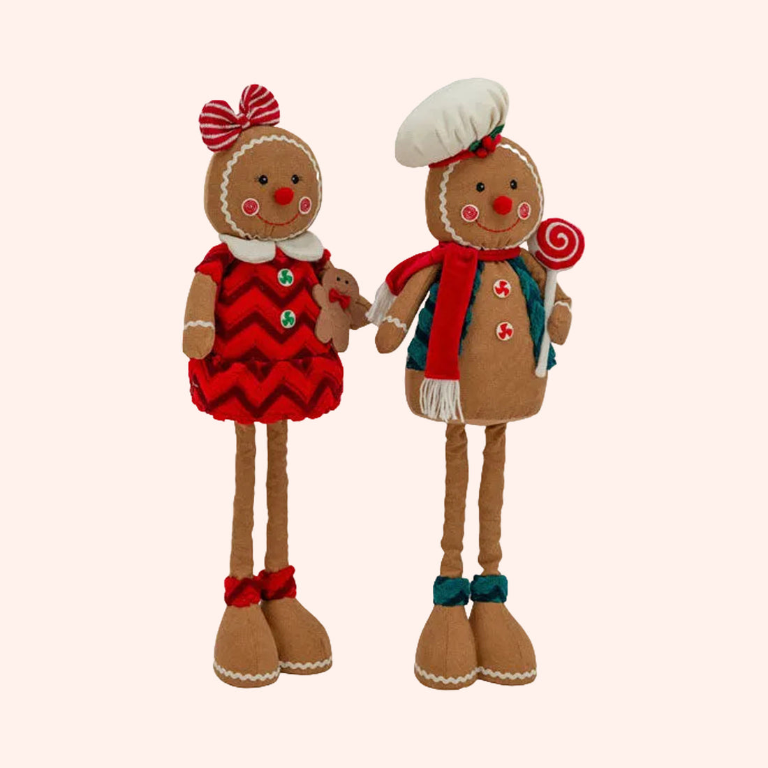 Two festive gingerbread-themed plush dolls standing side by side against a solid pale pink background. One doll wears a red chevron dress with a red bow on its head, and the other wears a white chef's hat, a red scarf, and a teal apron while holding a red-and-white lollipop.