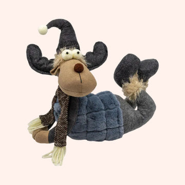 A plush stuffed animal shaped like a moose lying down, wearing a dark gray pointed hat with a white pom-pom, a brown patterned scarf, and a blue quilted vest. The moose has a tan face and neck, large white eyes, and fluffy beige hair, photographed on a plain background.