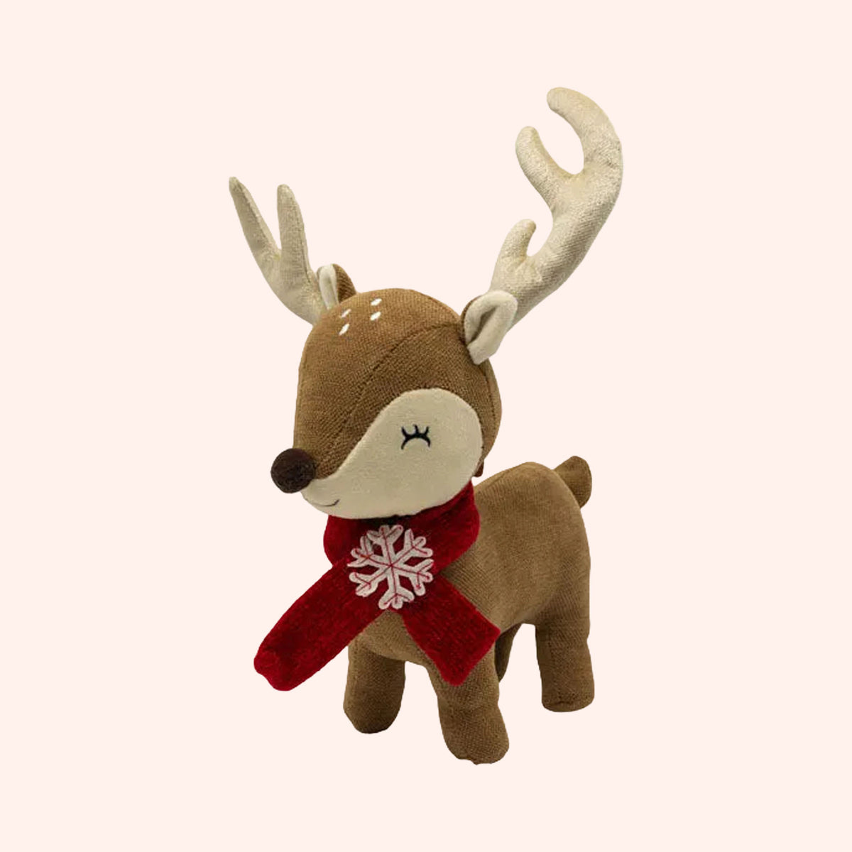 A plush toy reindeer with brown and cream fur, large cream antlers, and a red scarf with a white snowflake, shown on a solid pale pink background.